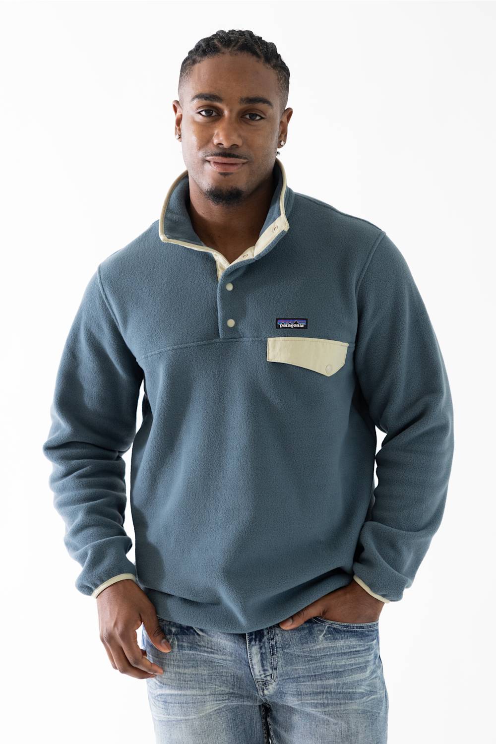A man wearing a Patagonia fleece pullover in slate blue with cream-colored trim on the collar, placket, and chest pocket, paired with blue jeans against a white studio background.