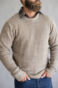 A man wearing a textured beige knit crewneck sweater layered over a dark plaid collared shirt and blue jeans, shown from the chin down against a neutral gray background.