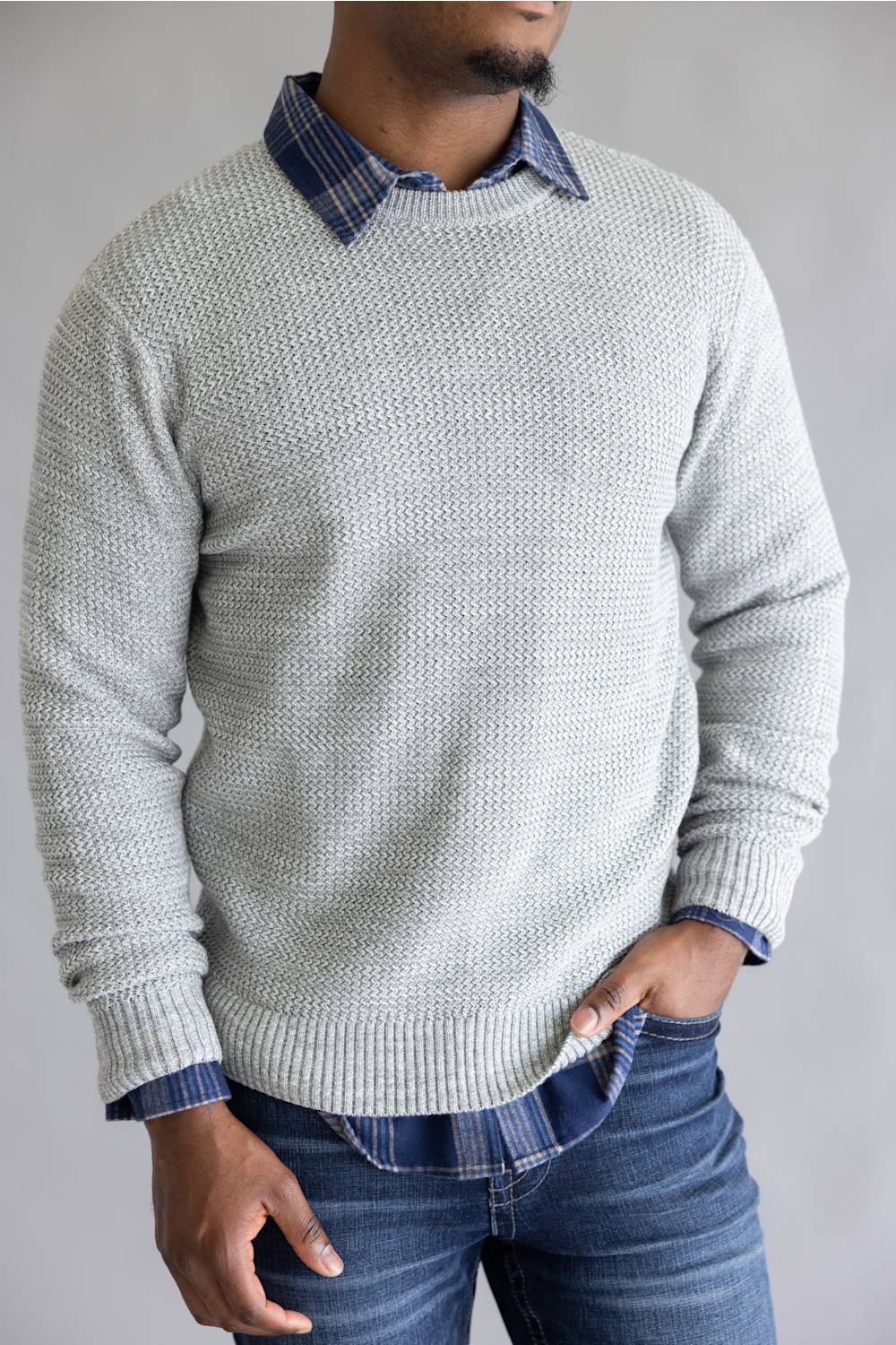 A man wearing a light gray textured knit sweater over a blue plaid collared shirt and blue jeans, shown from the waist up against a plain light gray background.