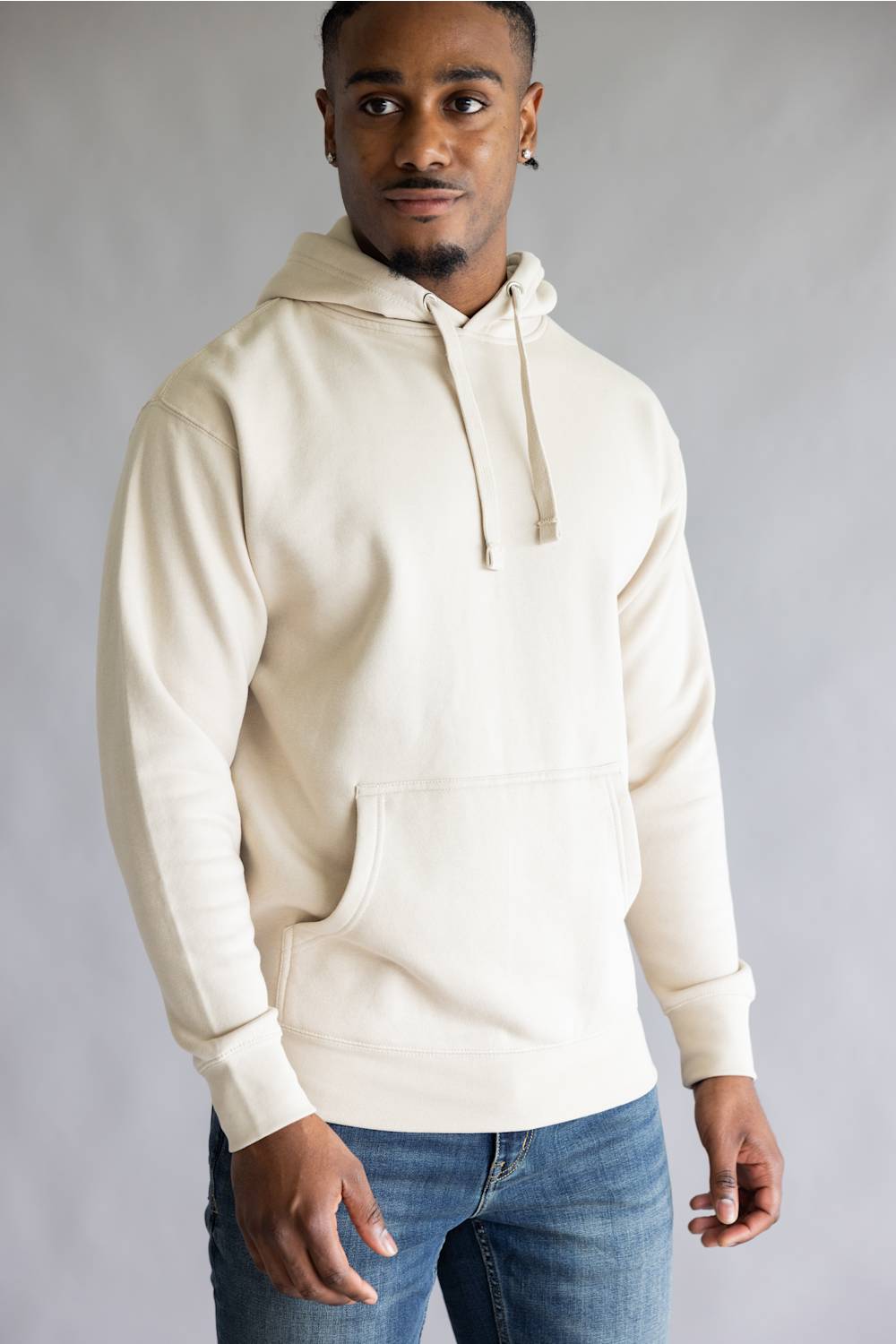 A man wearing a cream-colored pullover hoodie with drawstrings and a front kangaroo pocket, paired with blue jeans, standing against a neutral gray studio background.