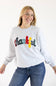 A smiling blonde woman wearing a light heather gray sweatshirt with the word 'thankful' written across the chest in multicolored letters, paired with blue jeans, against a plain white background.