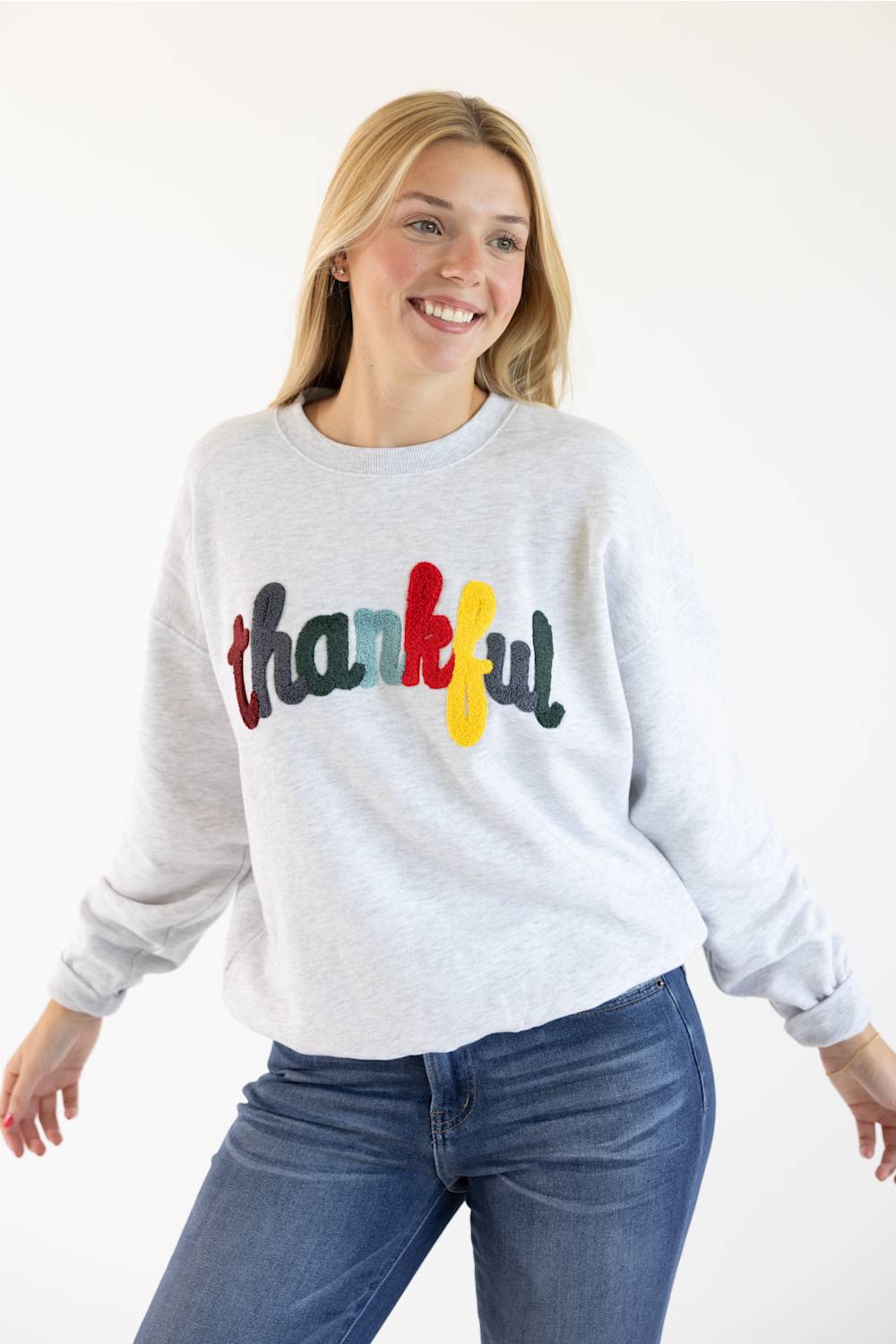 A smiling blonde woman wearing a light heather gray sweatshirt with the word 'thankful' written across the chest in multicolored letters, paired with blue jeans, against a plain white background.