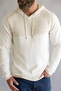 A man wearing a cream-colored knit hoodie with a textured, ribbed yoke across the chest and shoulders, paired with blue jeans against a neutral gray background.