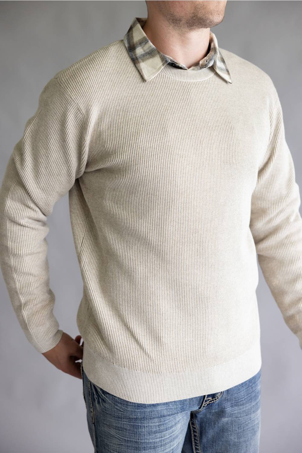 A man wearing a cream-colored ribbed knit sweater over a plaid collared shirt and blue jeans, photographed against a neutral gray background.