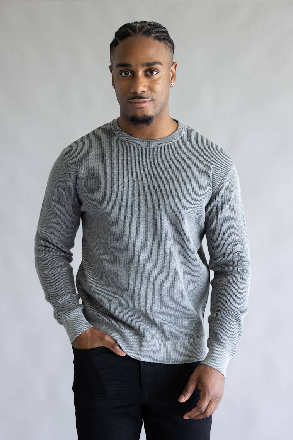 A man wearing a light gray crewneck knit sweater and black pants, standing against a neutral light gray background.