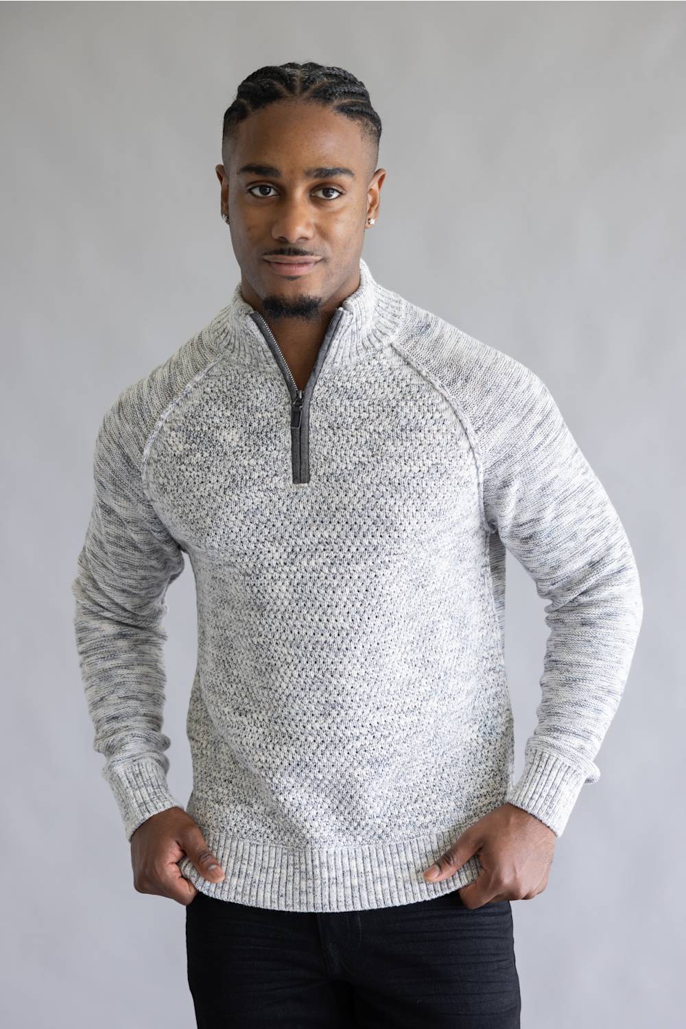 A man wearing a light gray and white marled knit quarter-zip sweater with dark pants, standing against a neutral gray studio background.