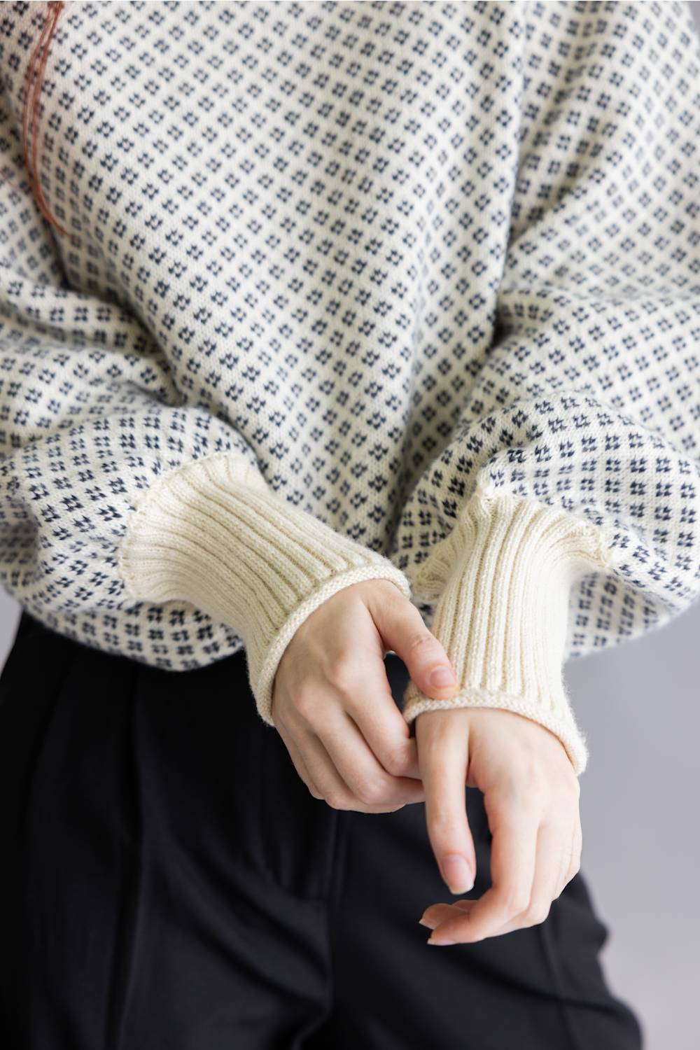 Close-up of a person wearing a cream ribbed knit sweater with a black and white micro-check pattern on the body and solid cream ribbed cuffs, paired with black pants.
