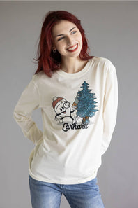 A smiling woman with red hair wearing a cream long-sleeve t-shirt with a cartoon graphic of a character next to a pine tree and the word 'Carhartt' printed below it, paired with blue jeans against a plain gray studio background.