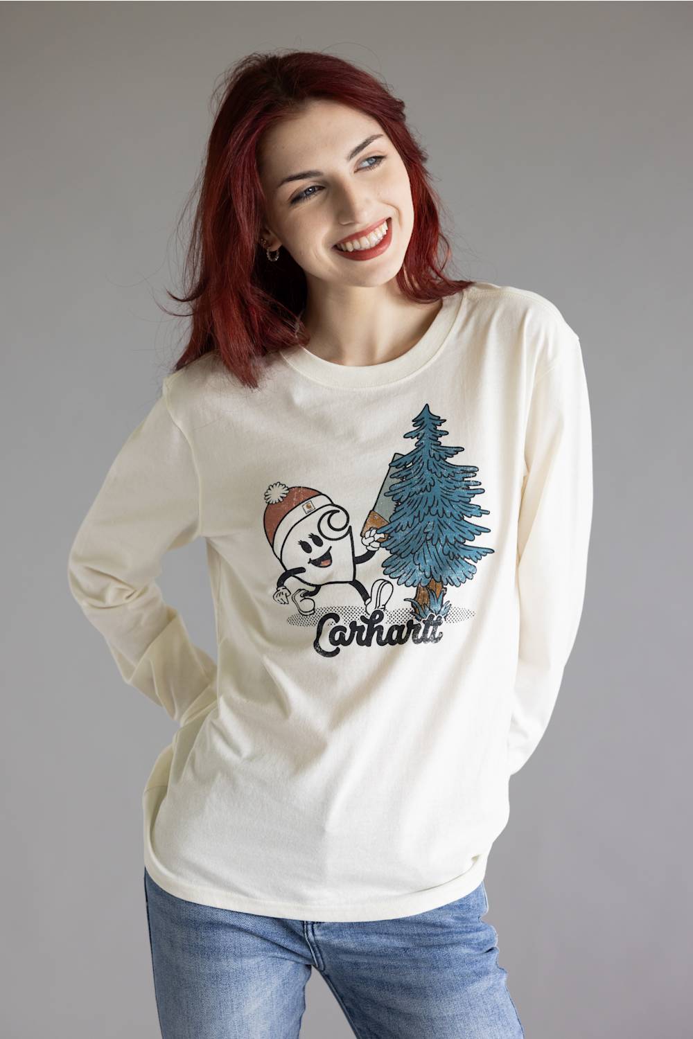 A smiling woman with red hair wearing a cream long-sleeve t-shirt with a cartoon graphic of a character next to a pine tree and the word 'Carhartt' printed below it, paired with blue jeans against a plain gray studio background.