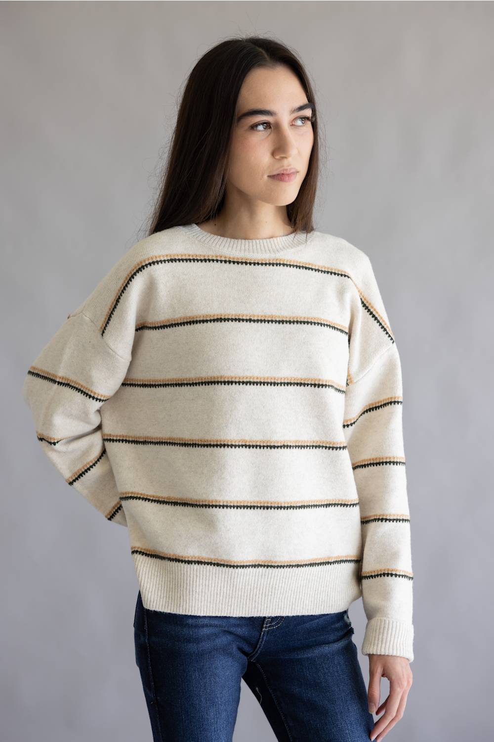 A woman wearing a cream-colored knit sweater with thin horizontal brown stripes and dark blue jeans, standing against a neutral gray background.