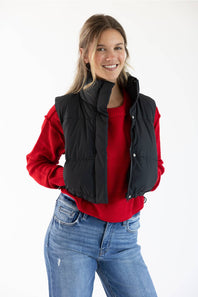 A woman wearing a black quilted puffer vest over a red long-sleeve top and blue jeans, photographed against a plain white studio background.