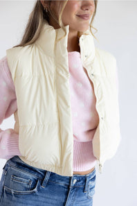 A woman wearing a cream-colored sleeveless puffer vest over a pink knit sweater and blue jeans, shown from the waist up against a plain light background.