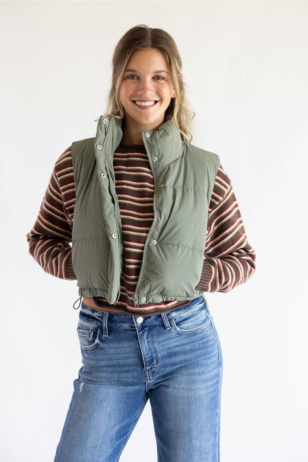 A smiling woman wearing an olive green puffer vest over a brown and red striped long-sleeve shirt, paired with blue jeans, against a plain white background.