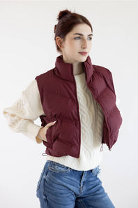 A woman wearing a burgundy quilted puffer vest over a cream cable-knit sweater and blue jeans, posing against a plain white studio background.
