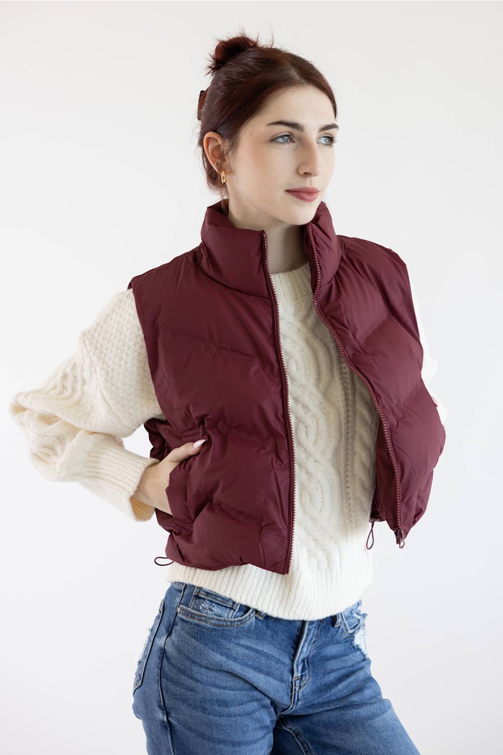 A woman wearing a burgundy quilted puffer vest over a cream cable-knit sweater and blue jeans, posing against a plain white studio background.