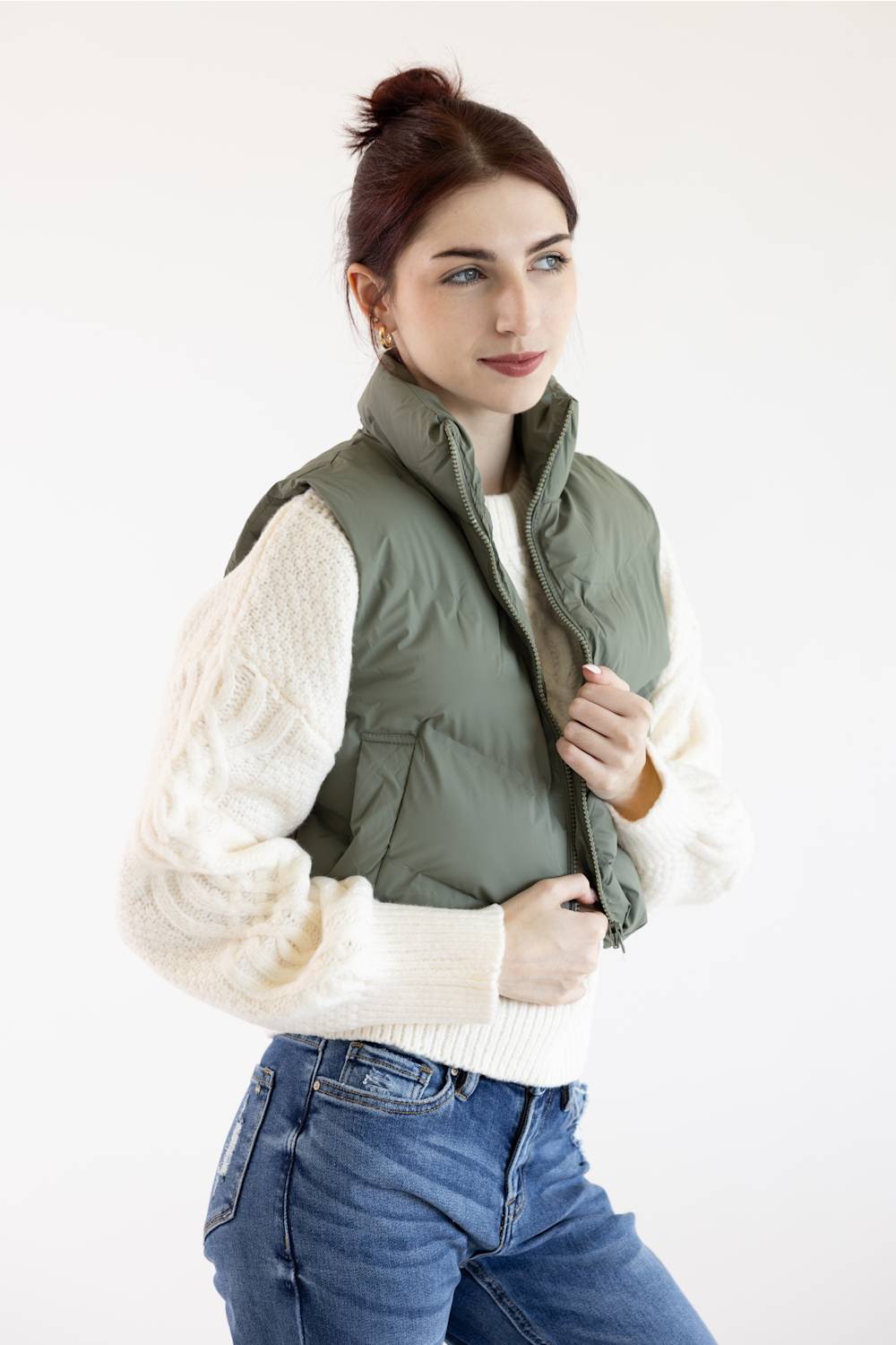 A woman wearing an olive green puffer vest over a cream cable-knit sweater and blue jeans, posing against a plain white studio background.