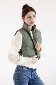 A woman wearing an olive green puffer vest over a cream cable-knit sweater and blue jeans, posing against a plain white studio background.