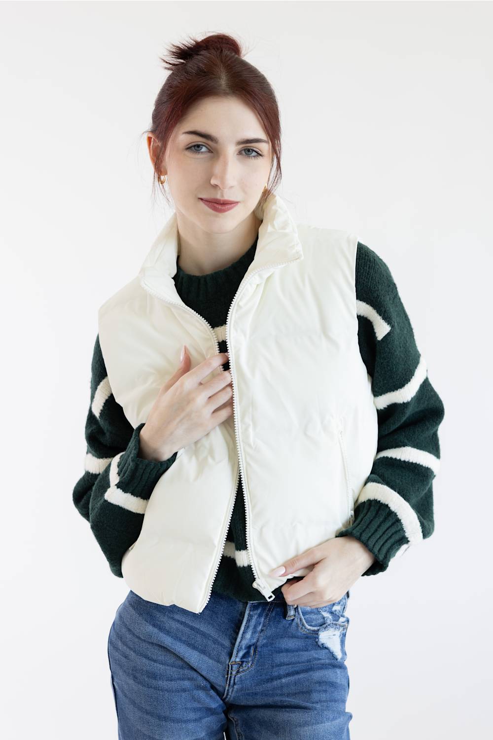 A woman wearing a cream-colored quilted puffer vest over a dark green and white striped knit sweater, paired with blue jeans, against a plain white studio background.