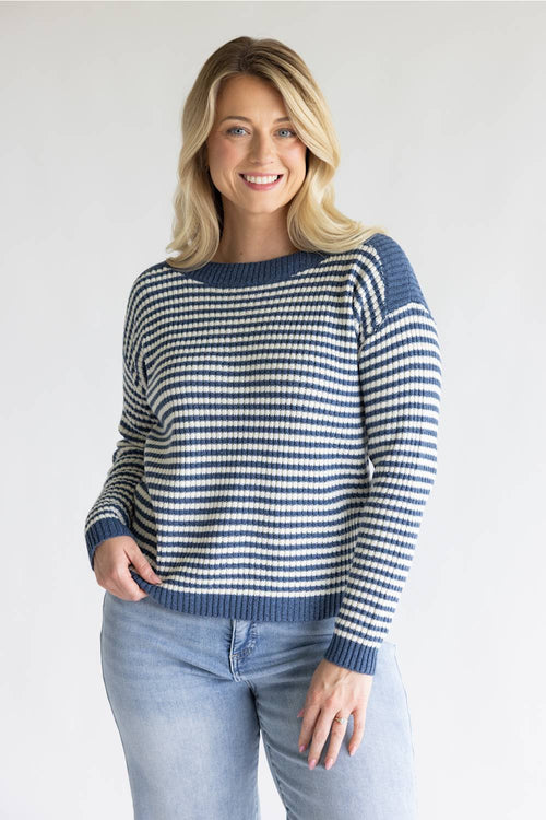 A woman wearing a navy and white striped knit sweater with blue jeans, standing against a light gray studio background.