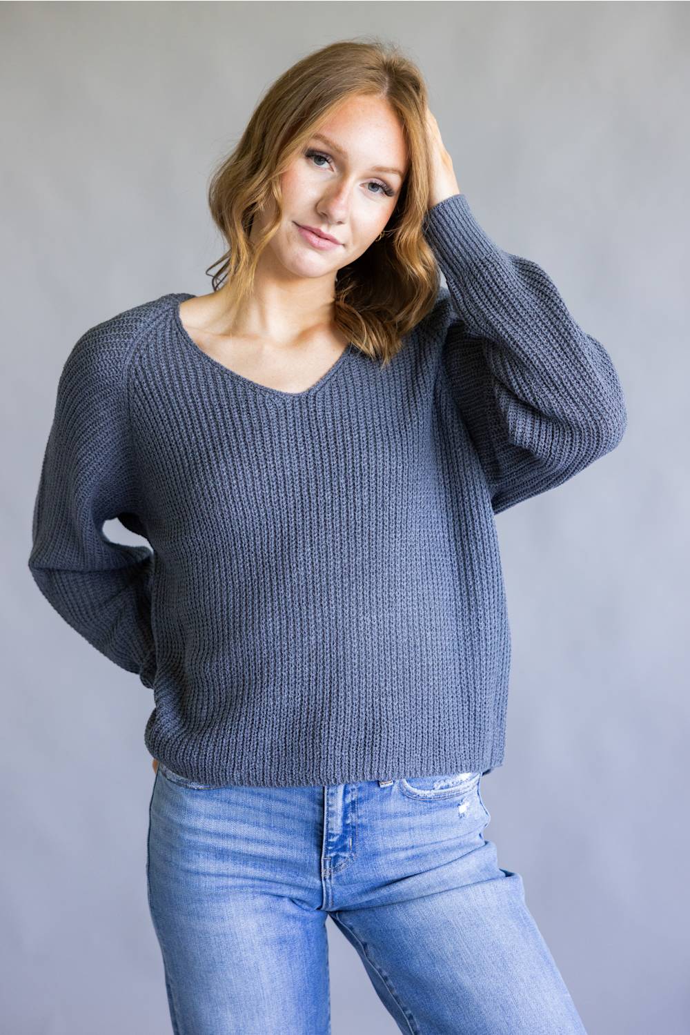 A woman wearing a slate blue ribbed knit sweater and light wash blue jeans, posing against a neutral gray studio background.