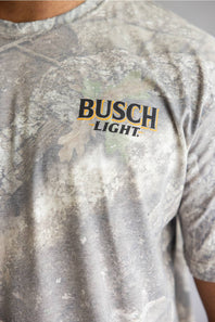 Close-up of a person wearing a Busch Light t-shirt with a gray and white tie-dye camouflage pattern and a black and gold logo on the chest.