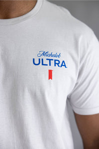 A close-up of a person wearing a white short-sleeve t-shirt with a blue 'Michelob ULTRA' logo and a small red ribbon graphic on the chest.