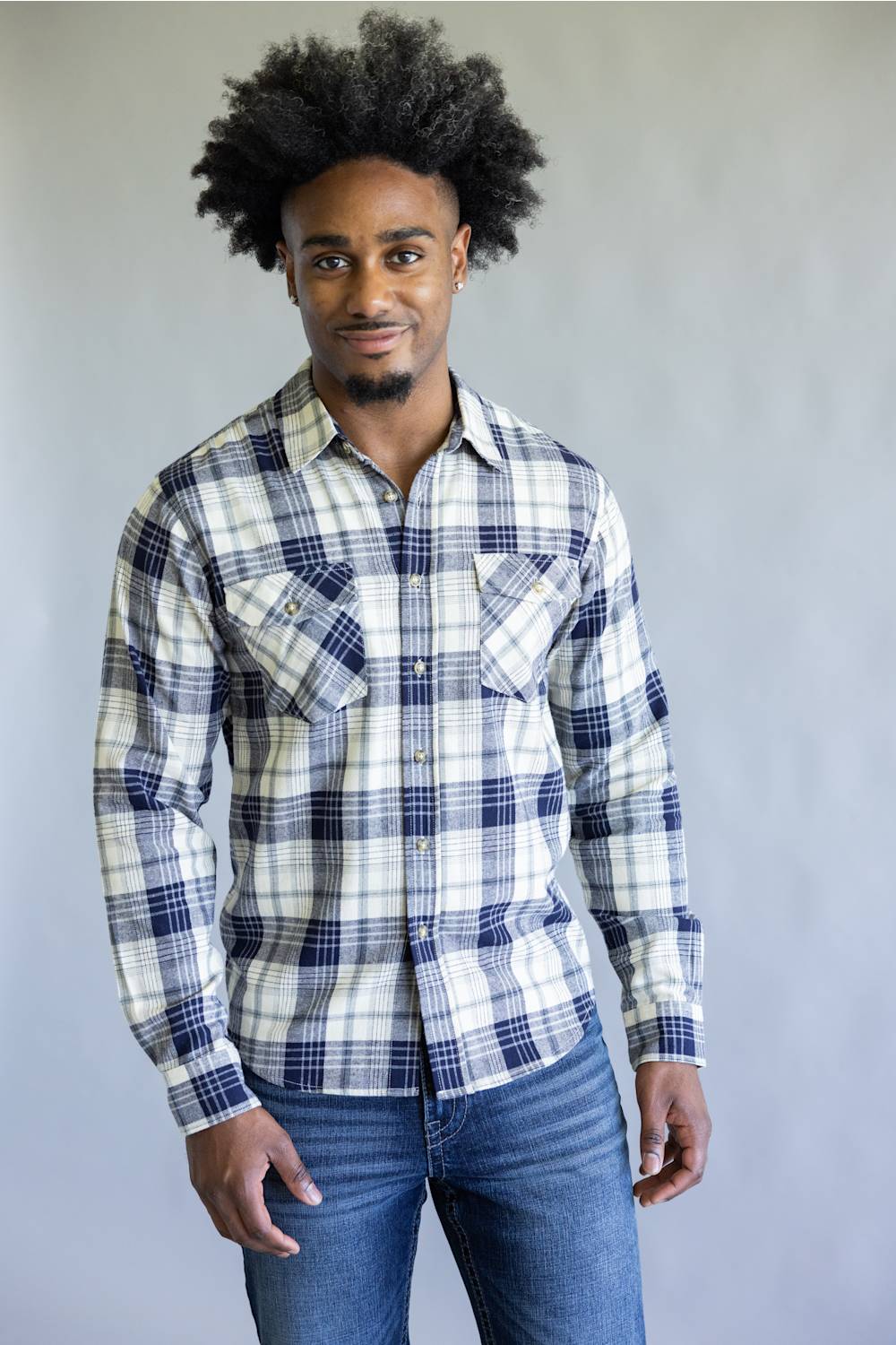 A man with an afro hairstyle wearing a long-sleeve plaid button-down shirt in white, navy, and gray, paired with blue jeans, standing against a plain gray studio background.