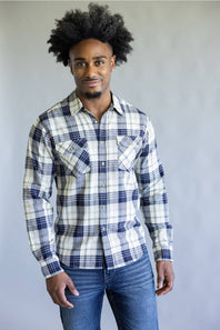 A man with an afro hairstyle wearing a long-sleeve plaid button-down shirt in white, navy, and gray, paired with blue jeans, standing against a plain gray studio background.