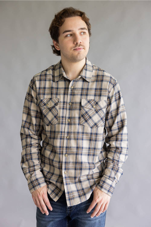 A man wearing a beige and navy plaid long-sleeve button-up shirt with two chest pockets, paired with blue jeans, standing against a neutral gray studio background.
