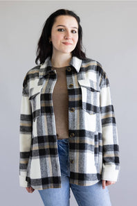 A woman wearing a black, white, and gray plaid button-up flannel shirt over a brown top and blue jeans, standing against a solid light gray background.
