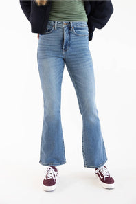 A person wearing light-wash blue flare jeans, a green top, a dark jacket, and burgundy sneakers, shown from the waist down against a plain white studio background.