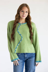 A woman modeling a light green knit sweater with a distinctive wavy teal trim and white bead details down the front, paired with blue jeans against a plain white background.
