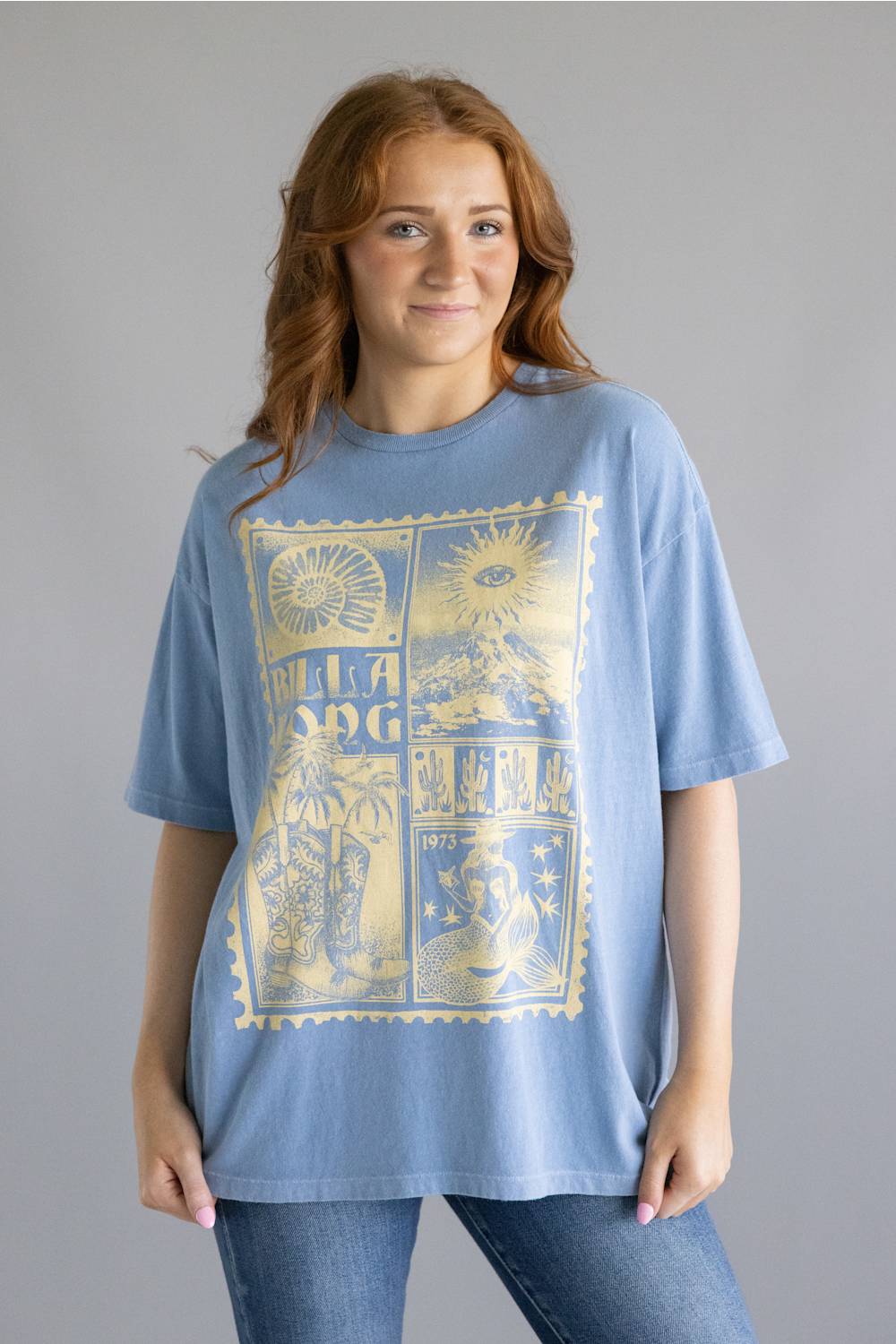 A woman wearing a light blue oversized graphic t-shirt with a large gold-toned vintage stamp design on the front, paired with blue jeans, against a solid gray studio background.