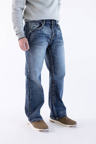 Men's medium wash straight-leg jeans featuring a side-profile view, subtle whiskering, and pairing with a grey crewneck sweater and brown casual shoes.