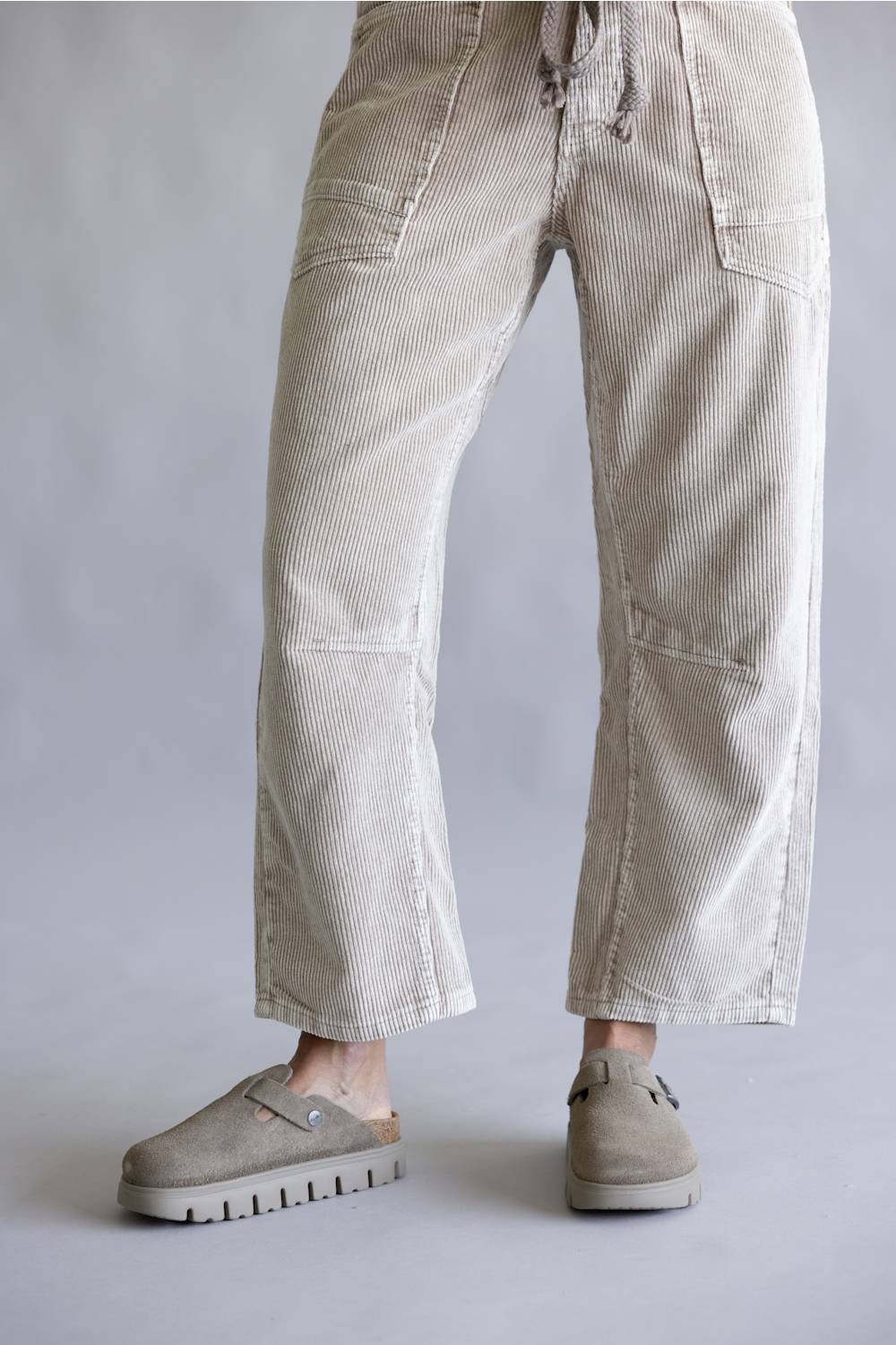 A person wearing light beige corduroy cropped pants with a drawstring waist and side pockets, paired with taupe slip-on shoes, photographed against a plain light gray studio background.