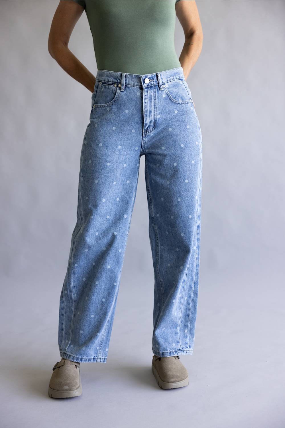 A person wearing light-wash blue denim jeans with a small white speckled pattern, styled with a sage green top and beige shoes against a neutral gray studio background.