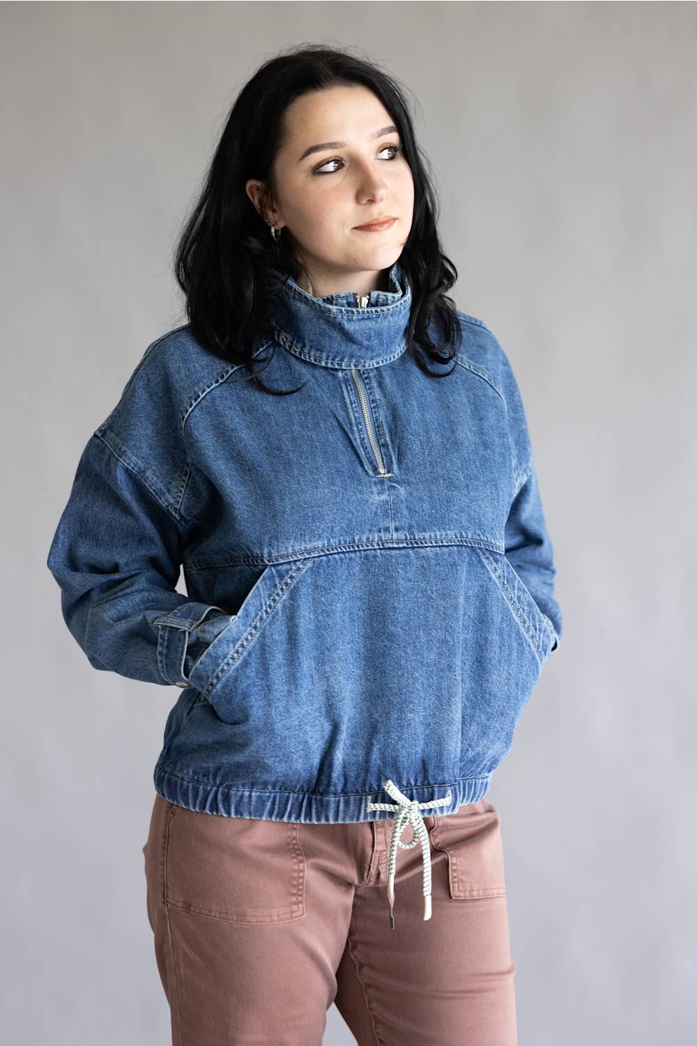 A woman wearing a blue denim half-zip pullover sweater with a mock neck and kangaroo pocket, paired with dusty rose drawstring pants, standing against a neutral gray studio background.