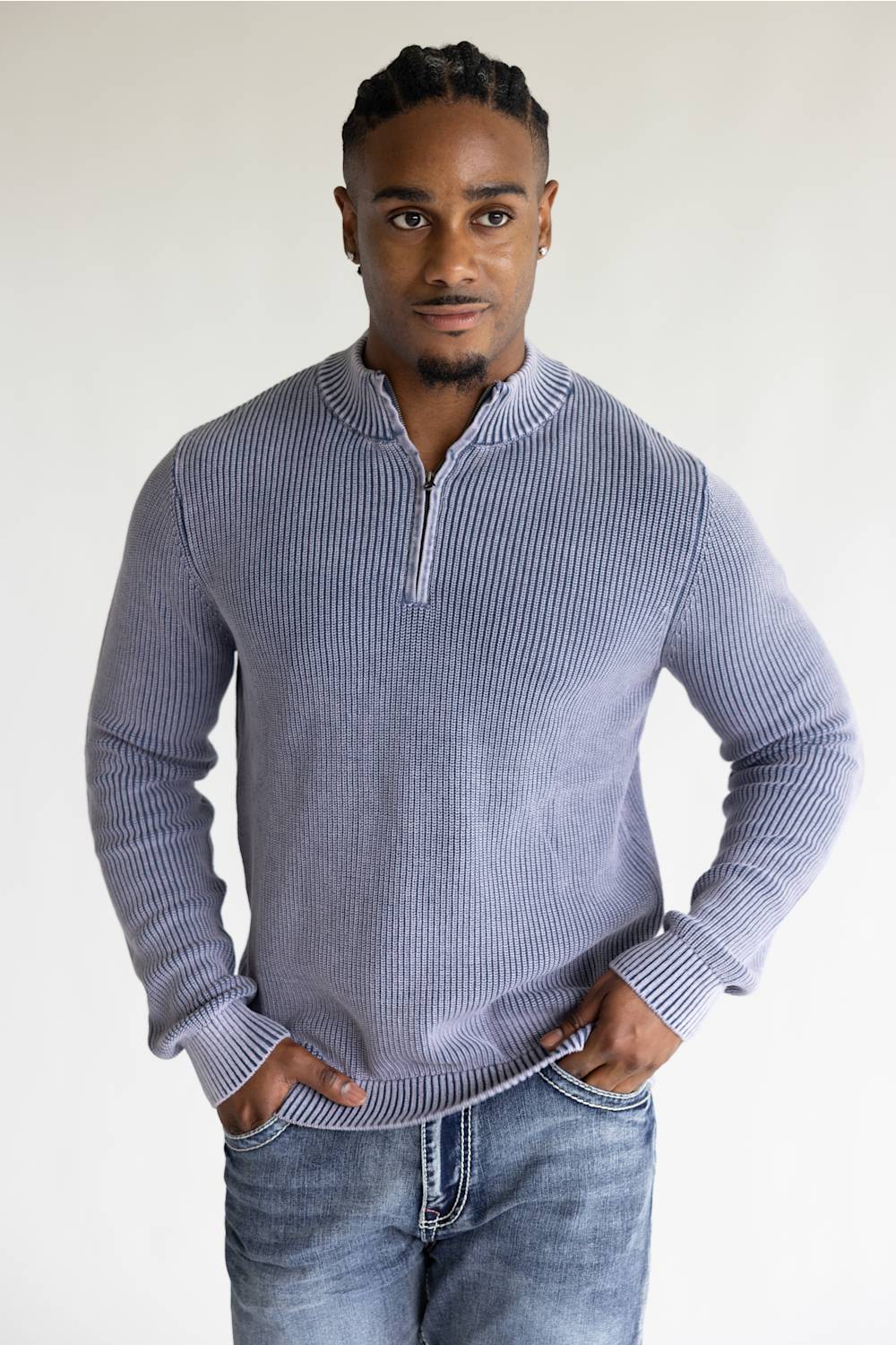A man wearing a ribbed long-sleeve half-zip sweater in a dusty lavender-gray color, paired with blue jeans, standing against a plain light background.