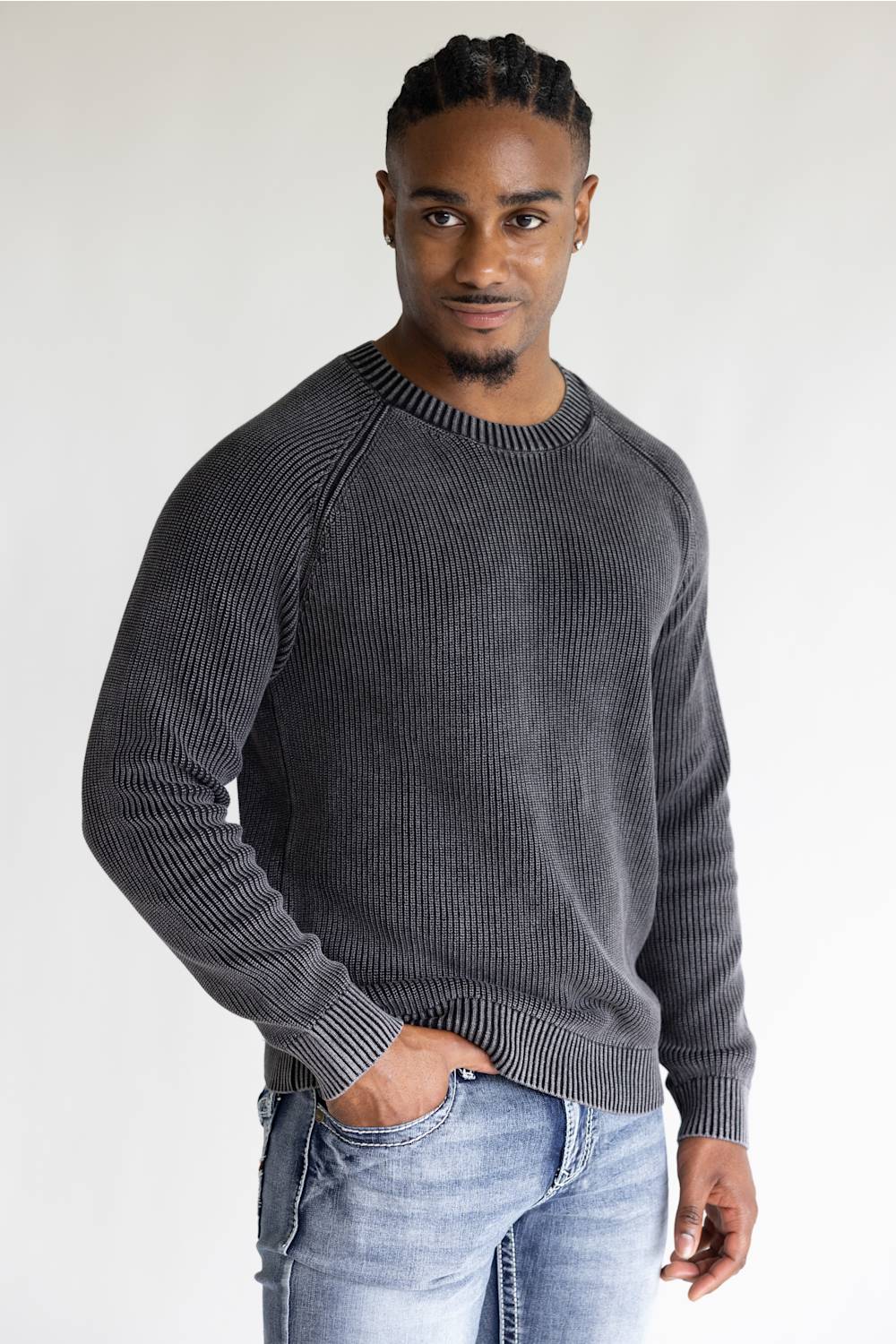 A man wearing a dark gray ribbed knit crewneck sweater and light blue jeans, standing against a plain light gray background.