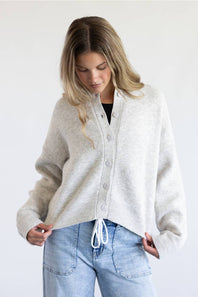 A woman modeling a light gray knit cardigan with a button-up front and drawstring detail, paired with light blue jeans against a plain white background.