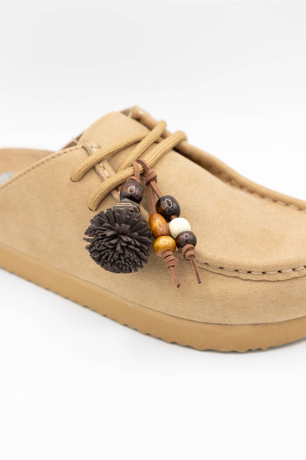A tan suede mule slipper with matching laces and a decorative hanging charm featuring wooden beads, a dark brown pom-pom, and a small white bead, shown on a white background.