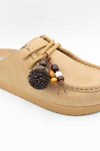 A tan suede mule slipper with matching laces and a decorative hanging charm featuring wooden beads, a dark brown pom-pom, and a small white bead, shown on a white background.