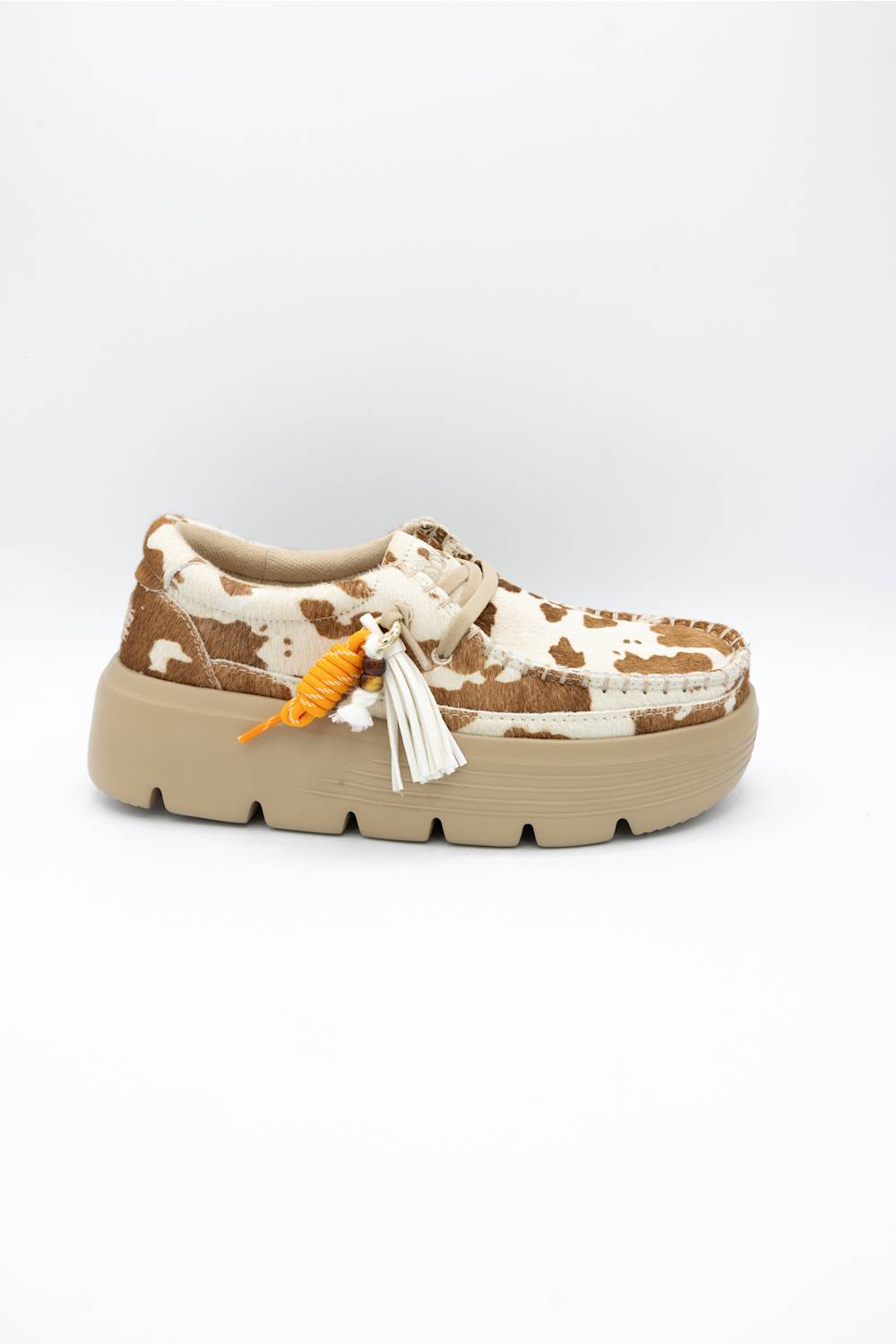 A single platform shoe with a brown and cream cow print upper, featuring a chunky beige sole, a white tassel, and an orange decorative accent.