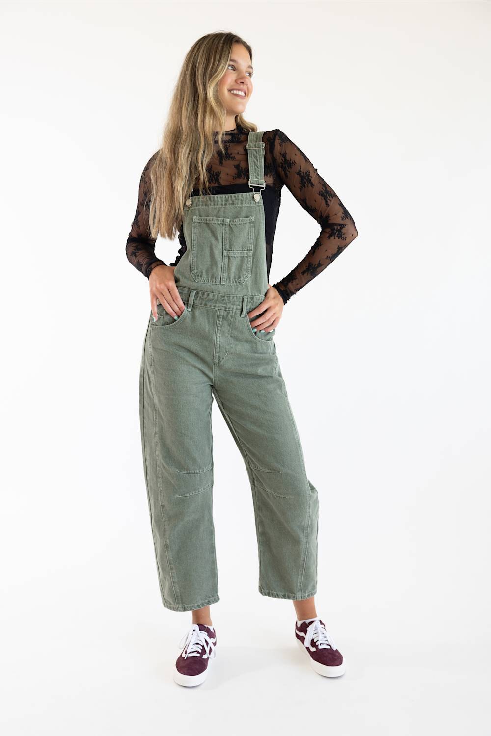 Front view of model wearing Aaron & Amber Slouchy Denim Overalls for Women in Olive featuring a relaxed barrel leg fit, classic chest bib pocket, and adjustable straps over a black patterned long-sleeve top paired with burgundy sneakers.