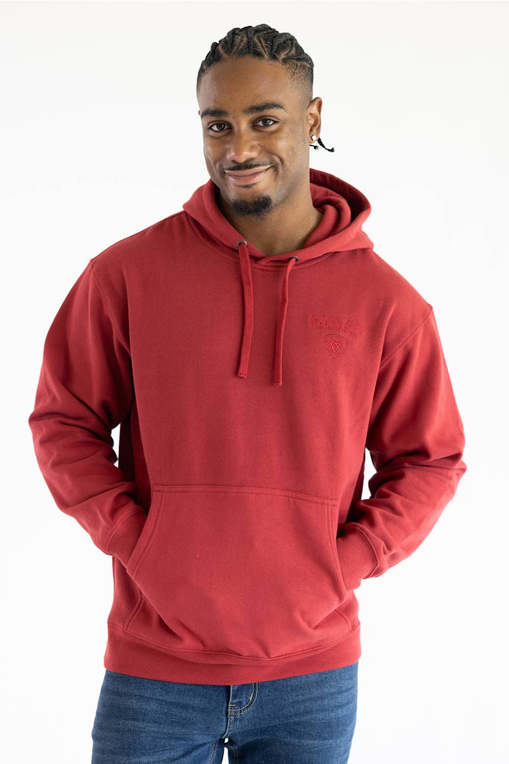 A man wearing a red pullover hoodie with a small tonal embroidered logo on the left chest, paired with blue jeans, photographed against a plain white background.