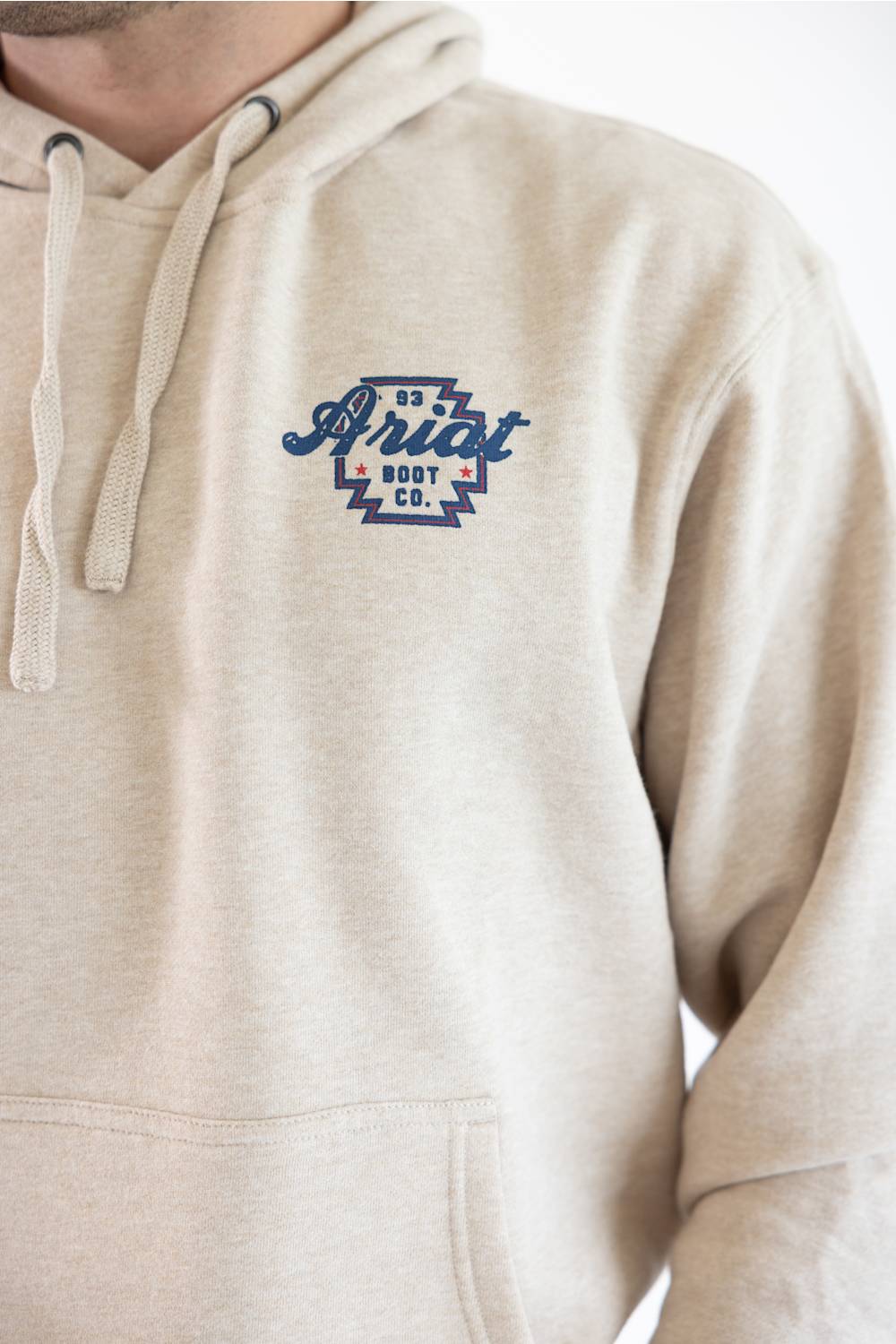 Close-up of a person wearing a light beige heather pullover hoodie with a blue and white embroidered chest logo that reads 'Ariat'.