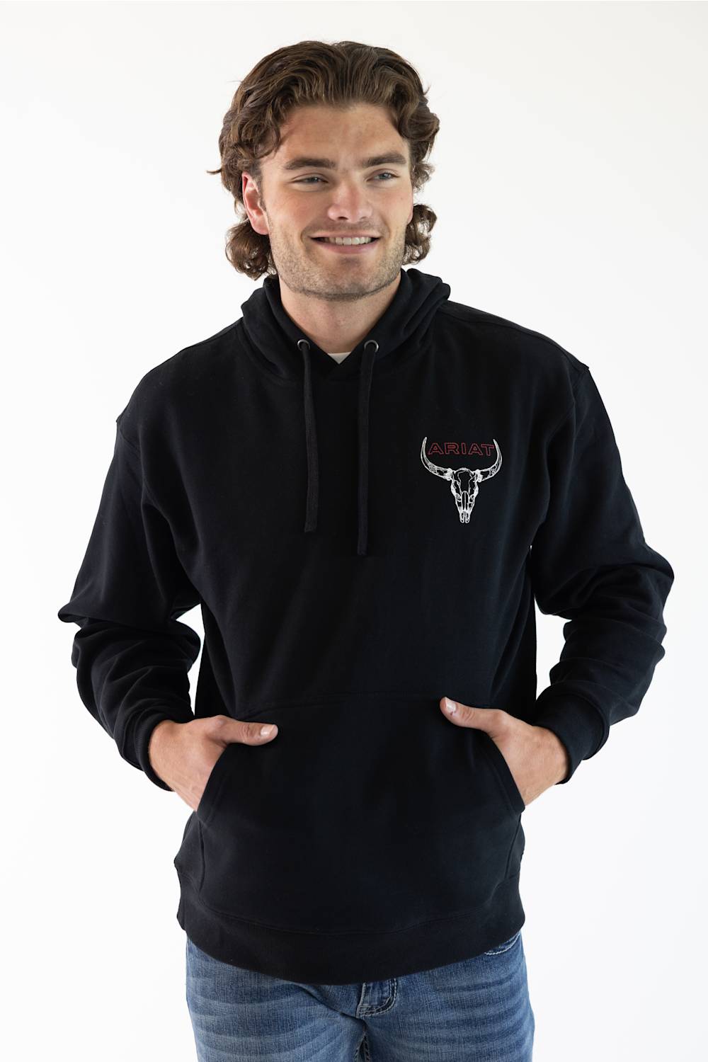 A man wearing a black pullover hoodie with a small bull skull logo on the left chest, paired with blue jeans, against a plain white background.