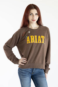 A woman with red hair wearing a brown crewneck sweatshirt with 'ARIAT' printed in yellow across the chest, paired with blue jeans, against a plain white background.