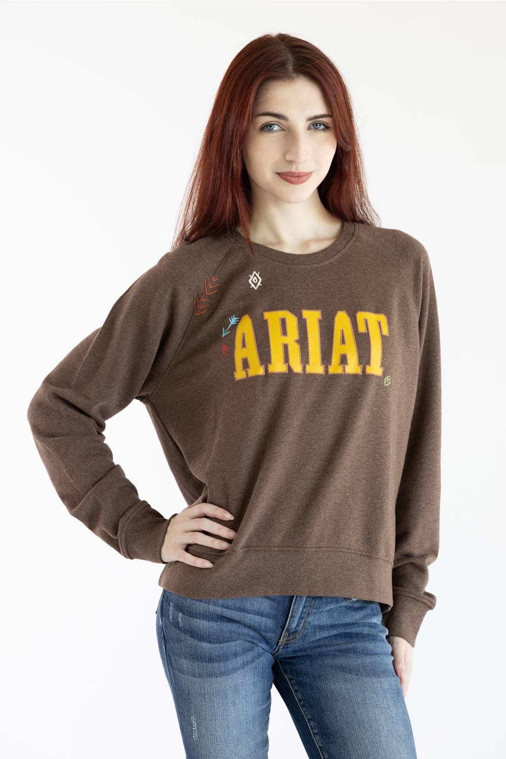 A woman with red hair wearing a brown crewneck sweatshirt with 'ARIAT' printed in yellow across the chest, paired with blue jeans, against a plain white background.
