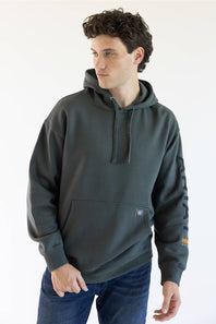 A man wearing a dark green hooded sweatshirt with a kangaroo pocket and blue jeans, standing against a plain white background.